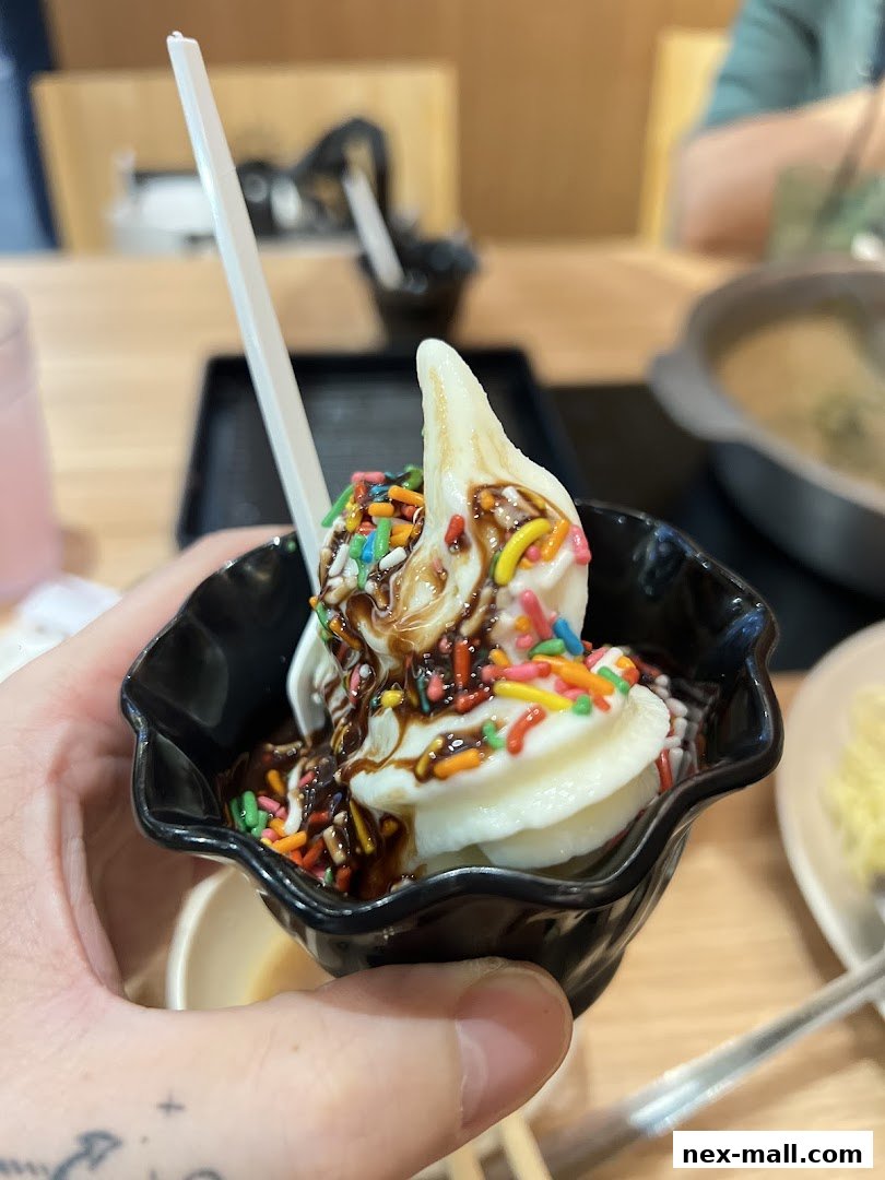 A hand holds a black bowl filled with soft-serve ice cream, chocolate sauce, and colorful sprinkles.