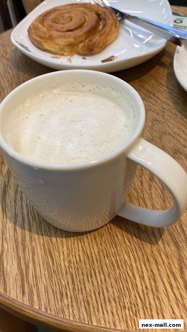 A close-up of a frothy white coffee drink in a white mug on a wooden table, with a pastry in the background.