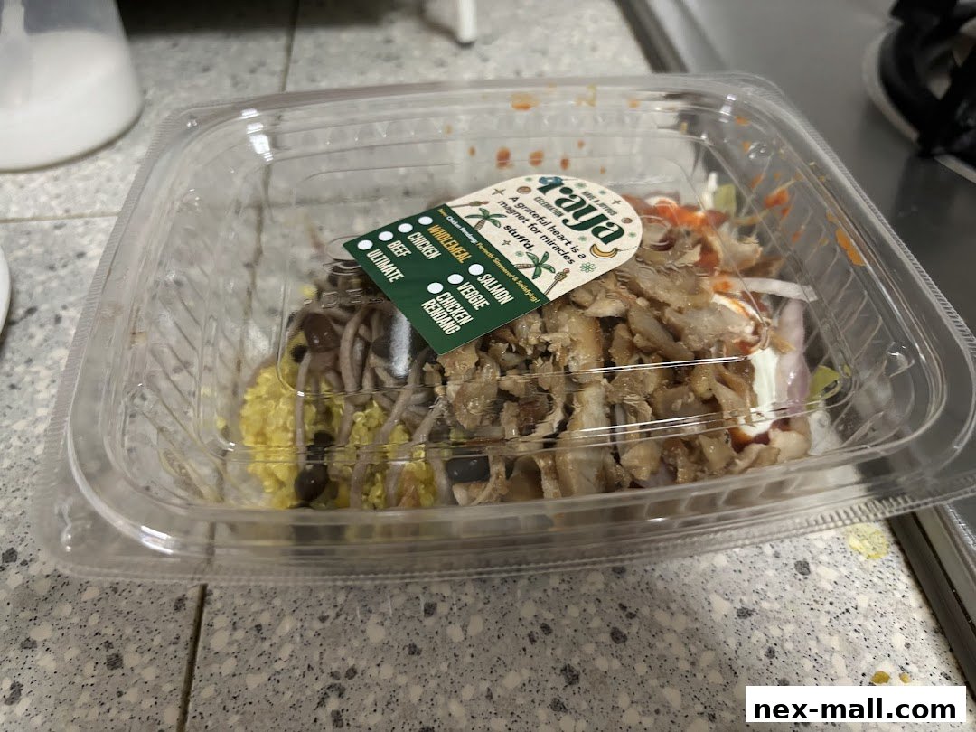 A clear plastic container filled with a meal, featuring various ingredients and a branded sticker.