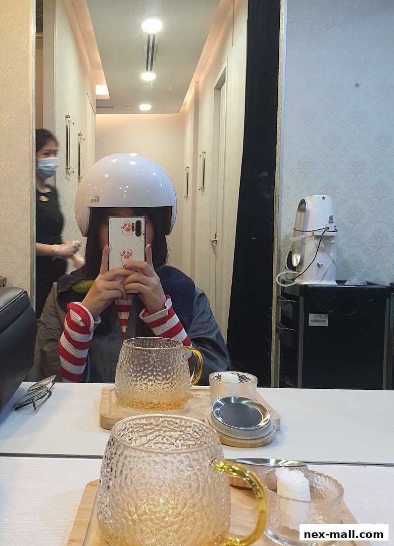 A person wearing a hair treatment helmet takes a selfie in a salon, with a staff member in the background.