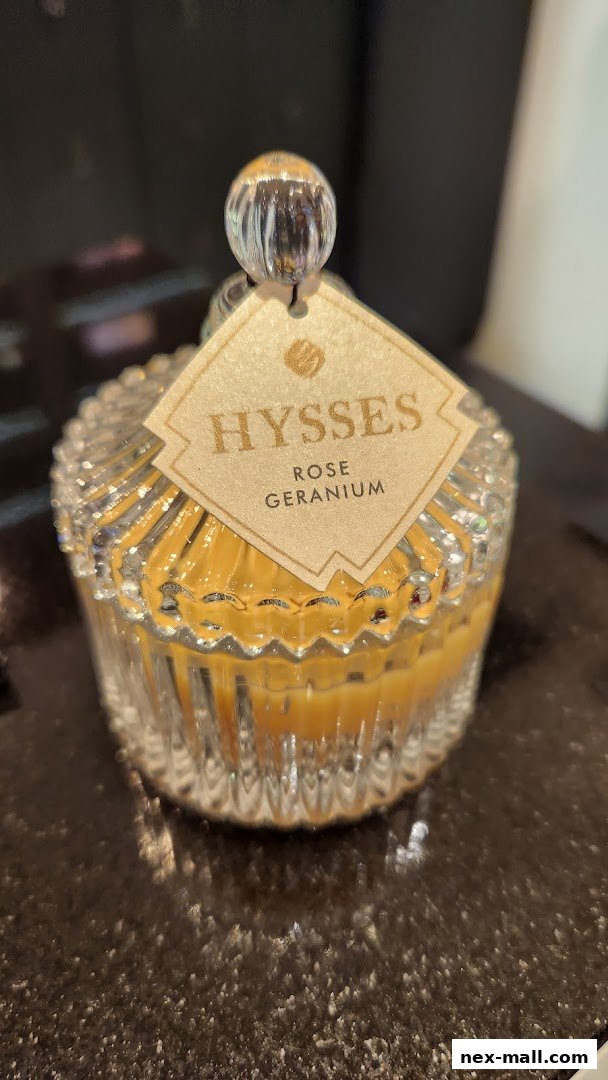 A close-up of a decorative glass jar candle with a 'HYSSES ROSE GERANIUM' label.