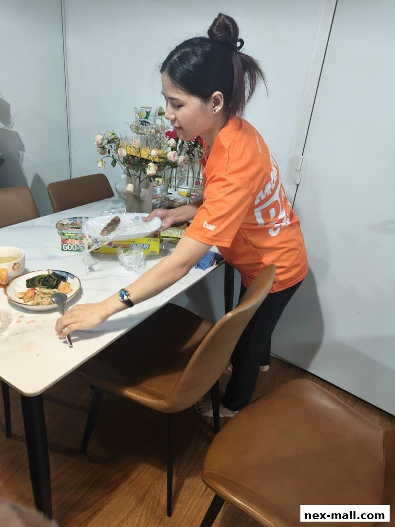 A woman in an orange t-shirt is clearing a table with food and flowers.