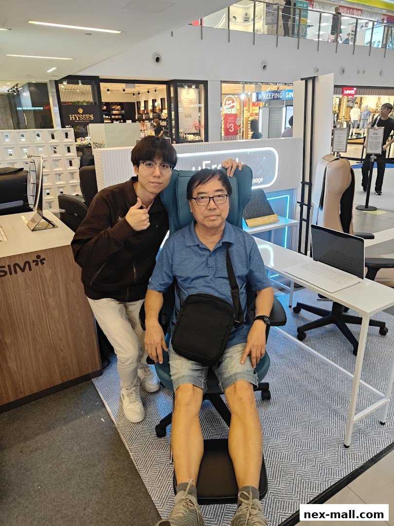 Two men are posing in a retail store, one sitting in a massage chair and the other standing and giving a thumbs up.
