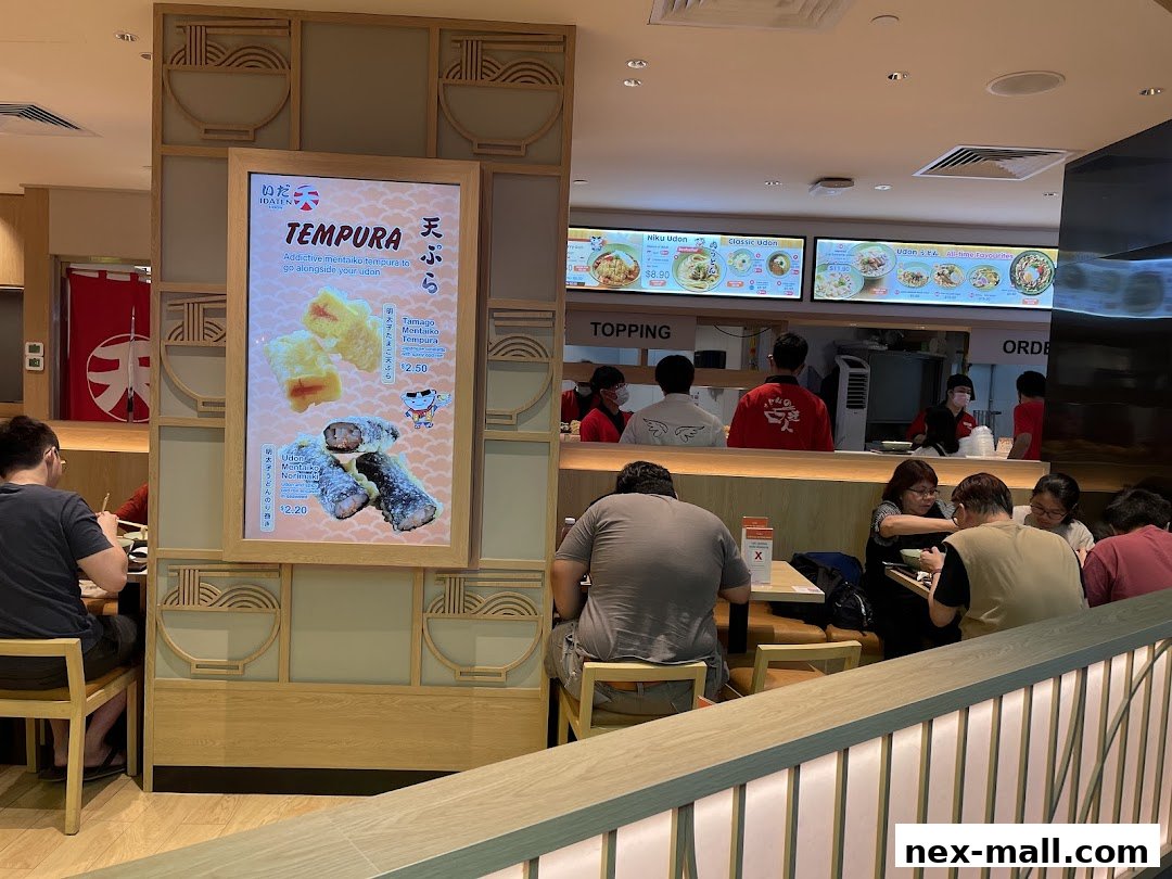 Diners are seated at tables in a Japanese udon restaurant with a prominent tempura menu display.