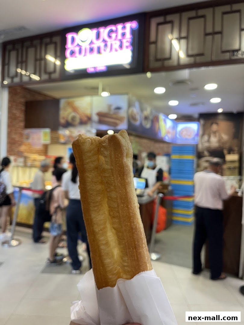 A person holds a golden-brown fried dough stick in front of a shop called Dough Culture.