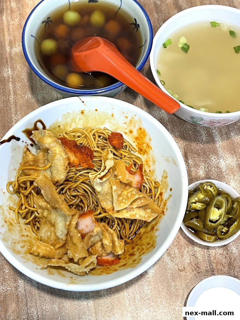 A bowl of noodles with char siu, fried wontons, and two side bowls of soup and pickled chilies.