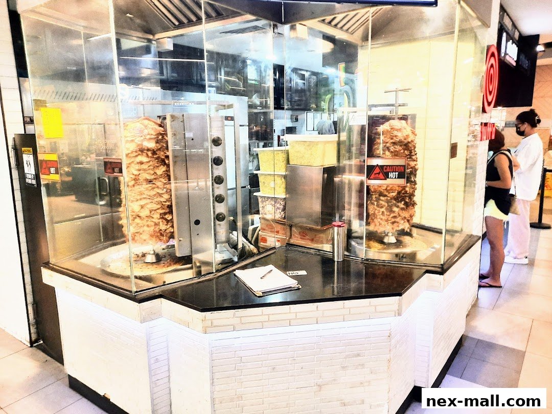 A food stall with rotating meat skewers and customers waiting in line.
