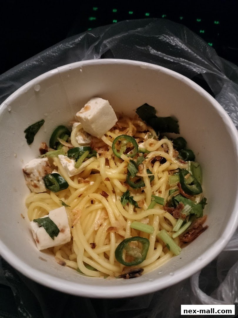 A close-up of a bowl of noodles with tofu, jalapeños, and herbs.
