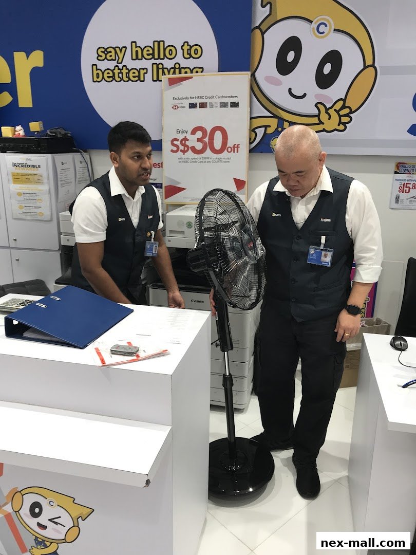 Two staff members stand near a fan and a counter with promotional materials.