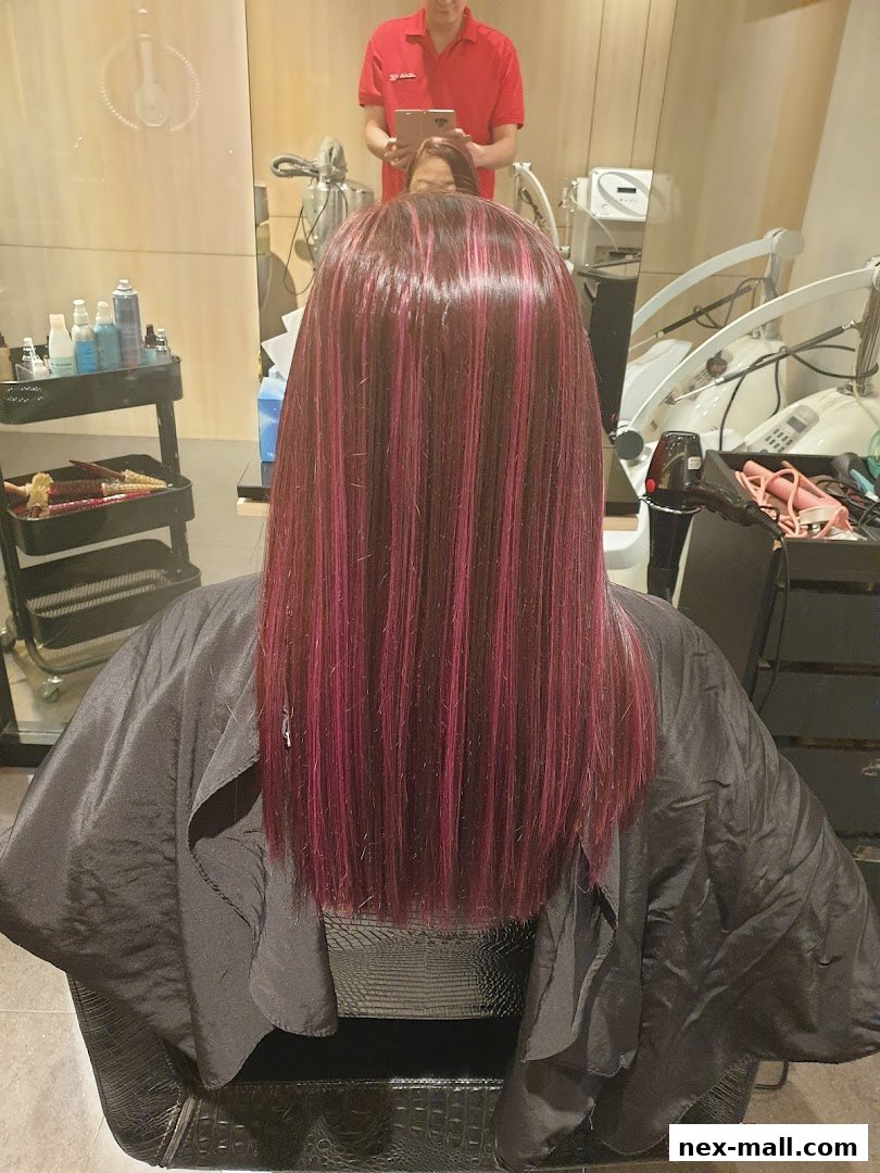 A person with long, dark hair with pink highlights sits in a salon chair, draped in a black cape.
