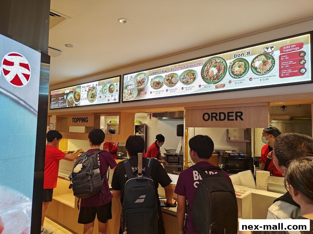 Customers queue to order at Idaten Udon, with a menu board displaying various udon and donburi dishes.