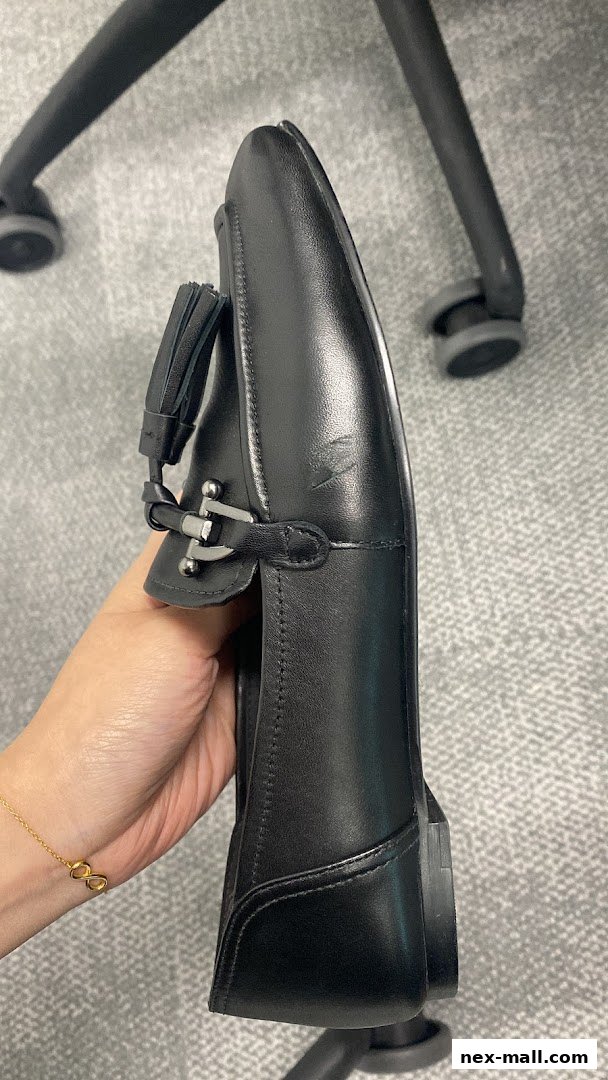 A hand holds a black leather loafer with a tassel and metal buckle detail.