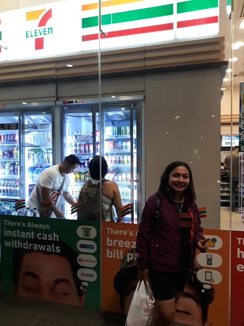 The exterior of a 7-Eleven store with customers and promotional signage for cash withdrawals and bill payments.