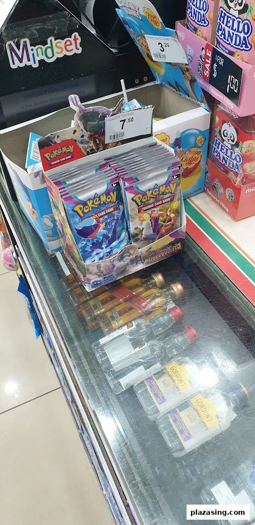 A retail counter display at 7-Eleven featuring Pokémon cards, popular snacks, and miniature alcohol bottles.