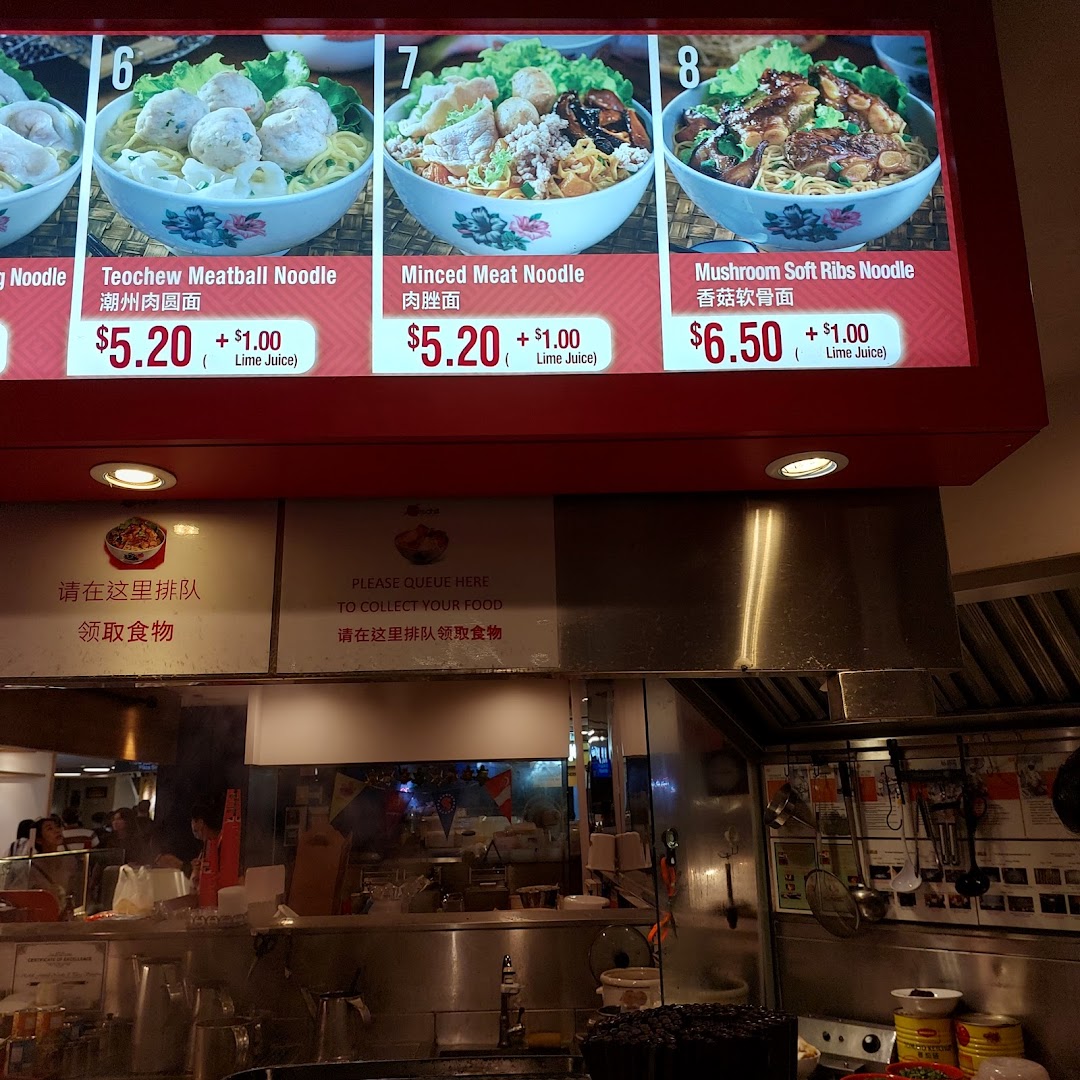 A digital menu board displays various noodle dishes with prices above a food stall counter.