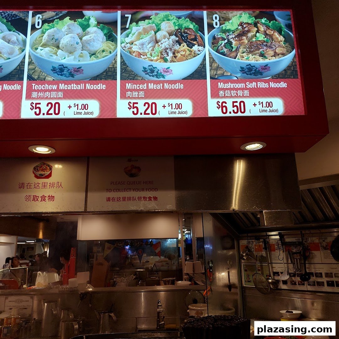 A digital menu board displays various noodle dishes with prices above a food stall counter.