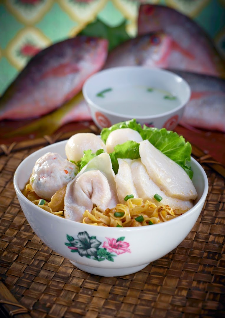 A vibrant bowl of Teochew fishball noodles with fish slices, fish dumplings, and a side of soup, with fresh fish in the background.