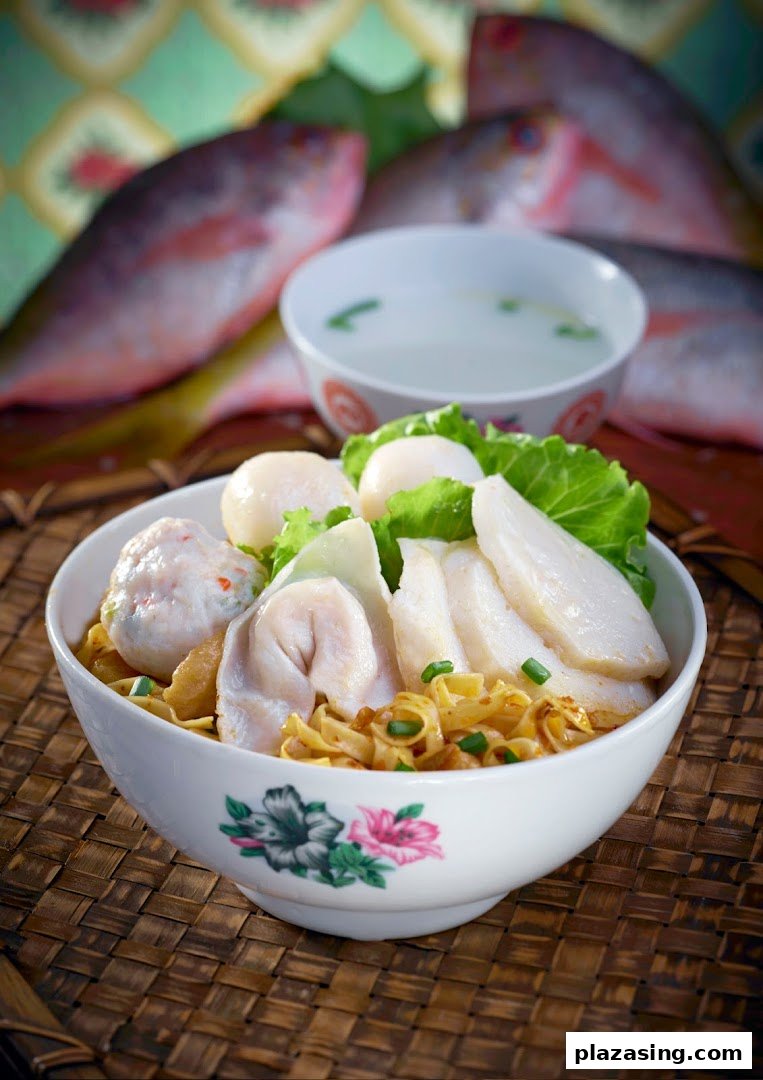 A vibrant bowl of Teochew fishball noodles with fish slices, fish dumplings, and a side of soup, with fresh fish in the background.
