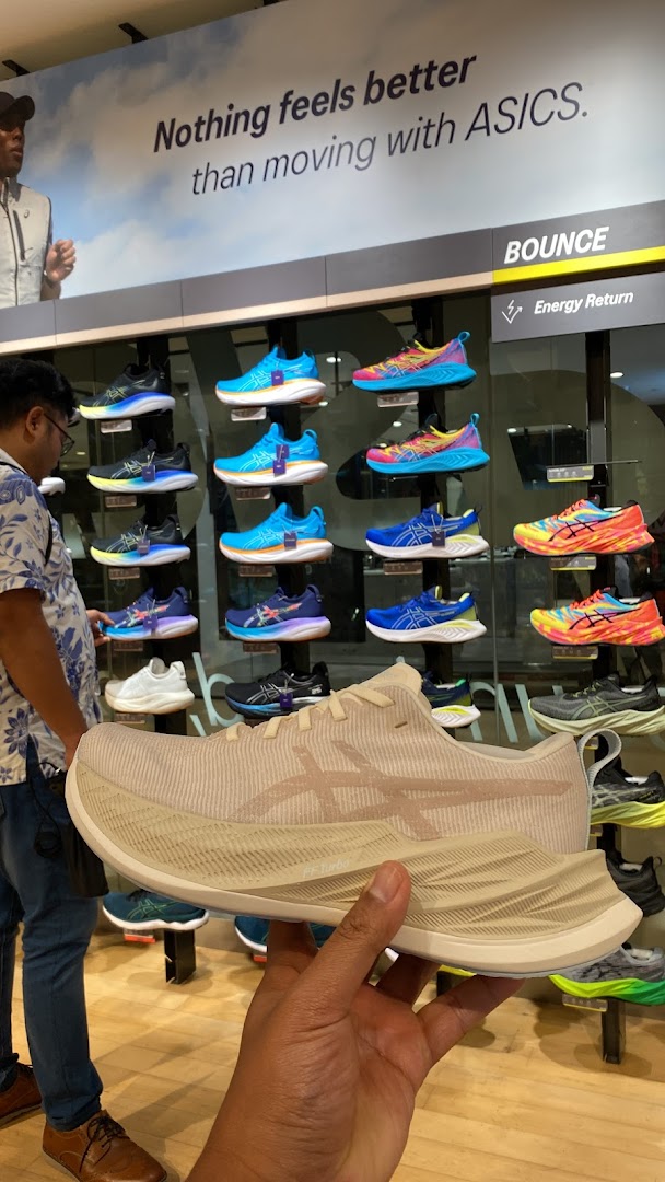 A person holds an ASICS running shoe in a store, surrounded by colorful displays of athletic footwear.