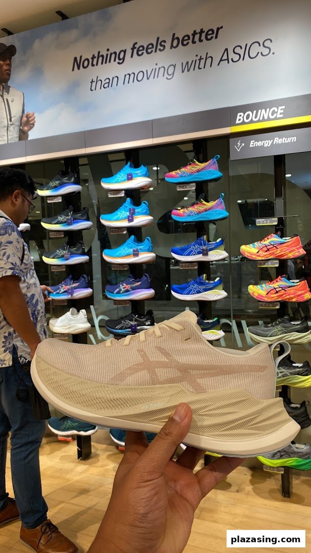 A person holds an ASICS running shoe in a store, surrounded by colorful displays of athletic footwear.