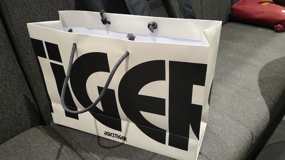A white ASICS TIGER shopping bag with black lettering and rope handles rests on a textured surface.