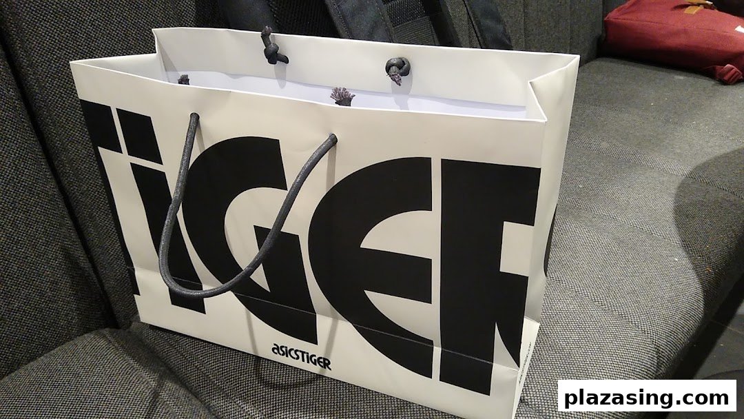 A white ASICS TIGER shopping bag with black lettering and rope handles rests on a textured surface.