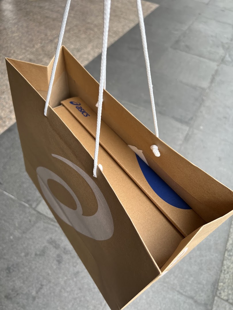 A brown ASICS shopping bag with white handles and logo, containing a product box.