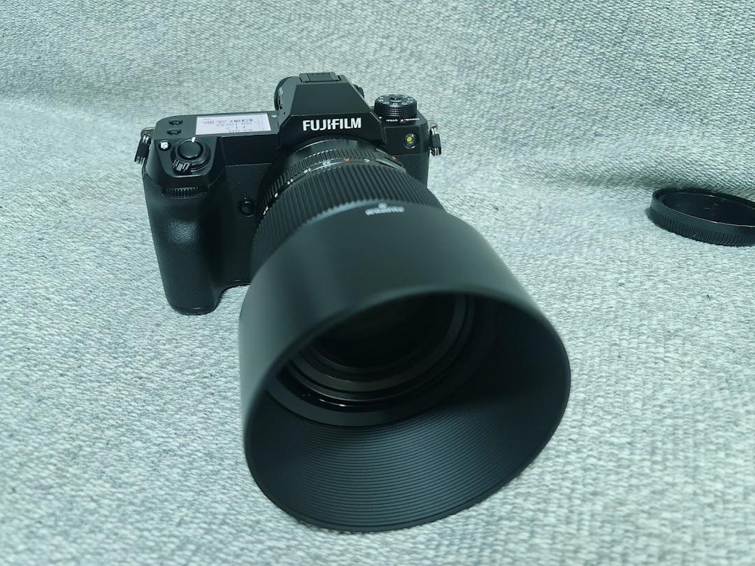 A black Fujifilm camera with a large lens and lens hood is displayed on a grey surface.