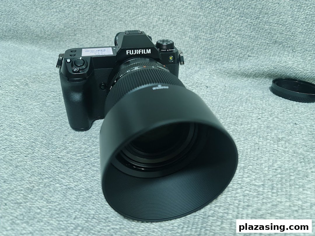A black Fujifilm camera with a large lens and lens hood is displayed on a grey surface.