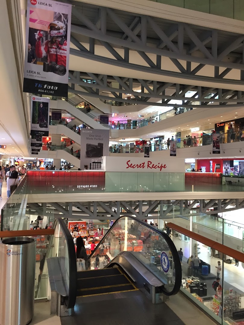 A vibrant multi-level shopping mall interior with escalators, various shops, and bustling activity.