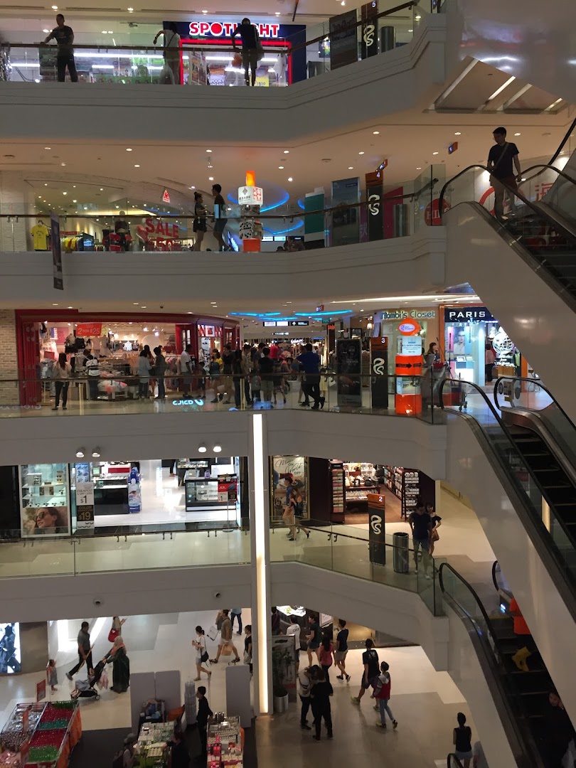 A bustling multi-level shopping mall interior with shoppers, escalators, and various retail stores.