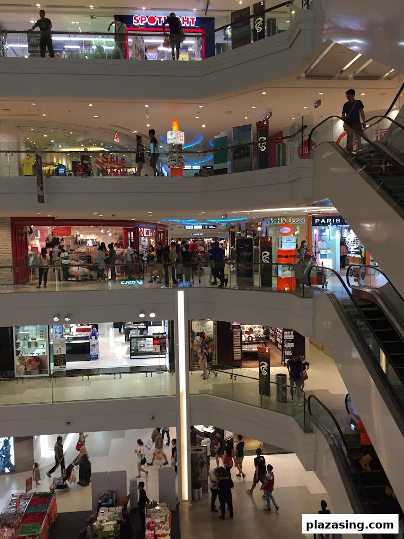 A bustling multi-level shopping mall interior with shoppers, escalators, and various retail stores.