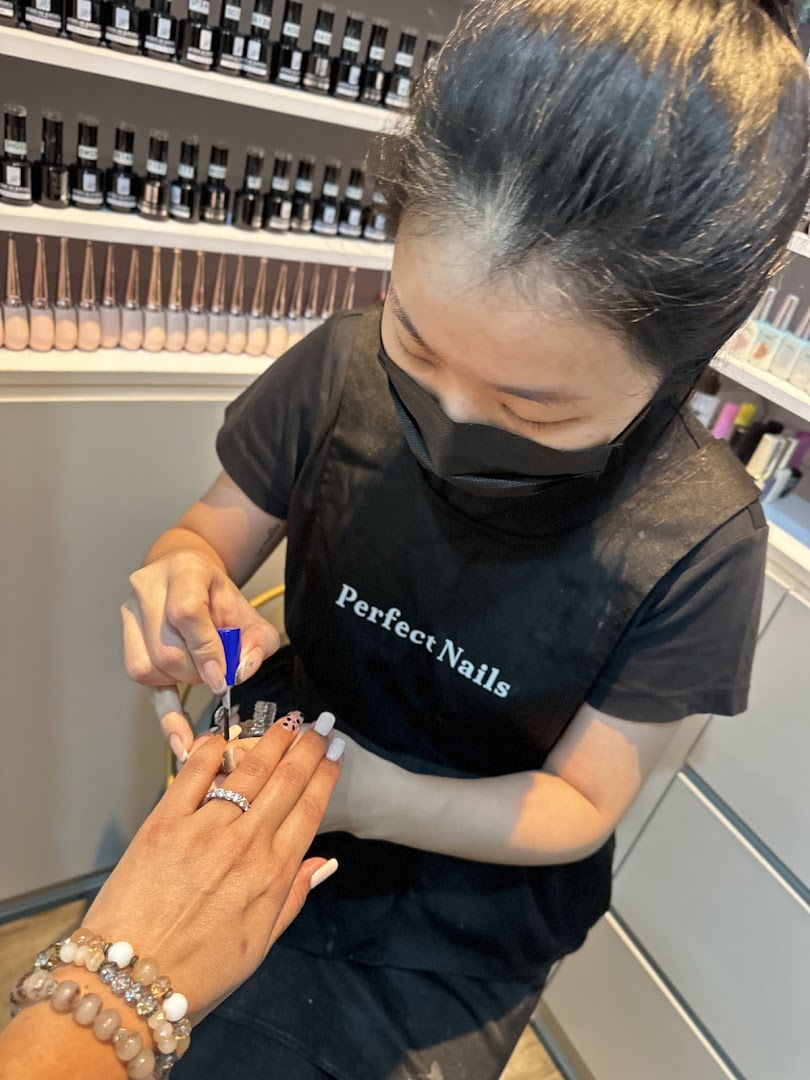 A nail technician, wearing a 'Perfect Nails' apron, meticulously applies polish to a customer's nails in a salon setting.