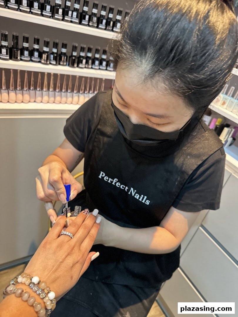 A nail technician, wearing a 'Perfect Nails' apron, meticulously applies polish to a customer's nails in a salon setting.