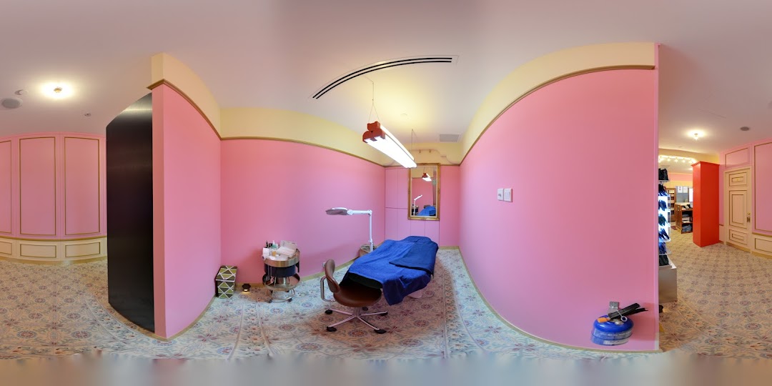 A pink-walled treatment room with a bed, chair, and beauty equipment, featuring ornate flooring.