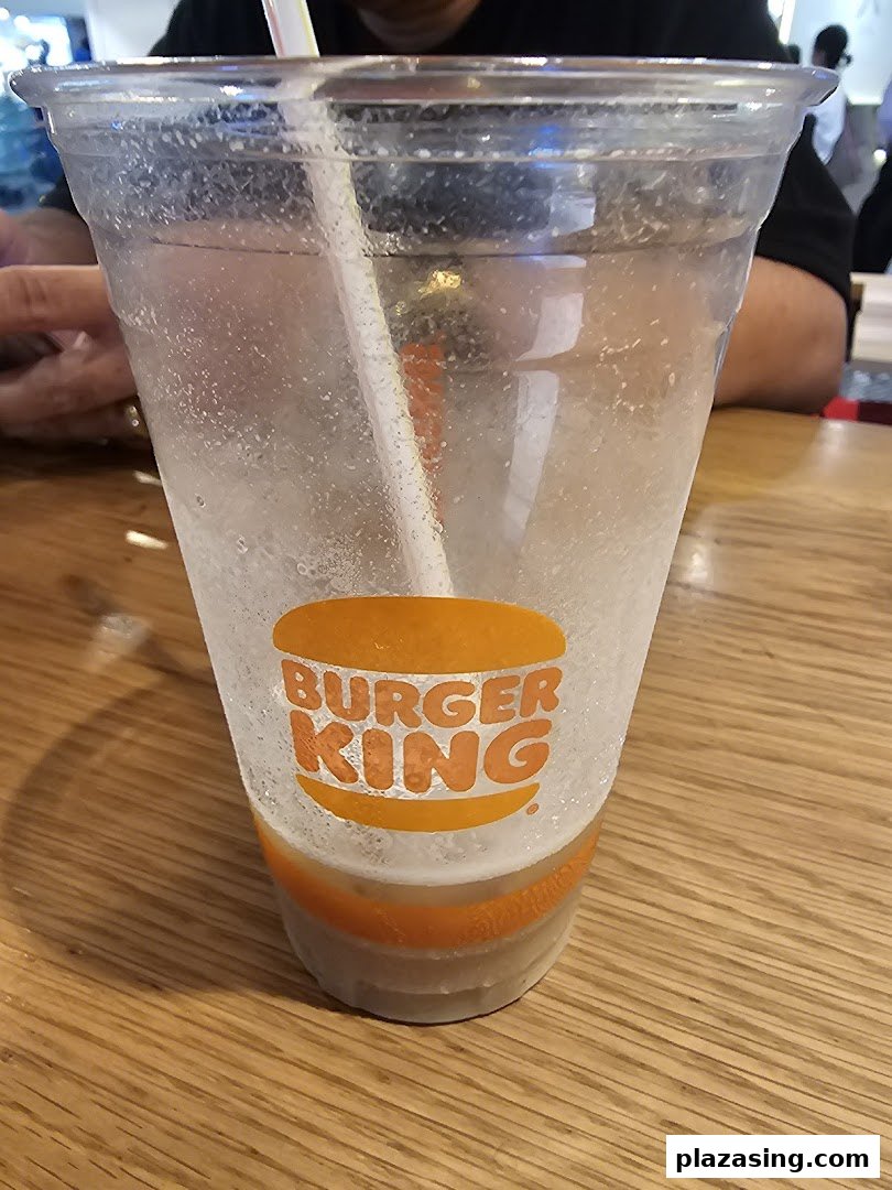 A clear plastic Burger King cup with a straw, partially filled, on a wooden table.