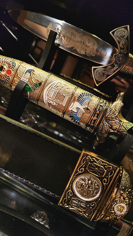 A close-up of a highly ornate, gold-colored decorative dagger with intricate historical and mythological designs.
