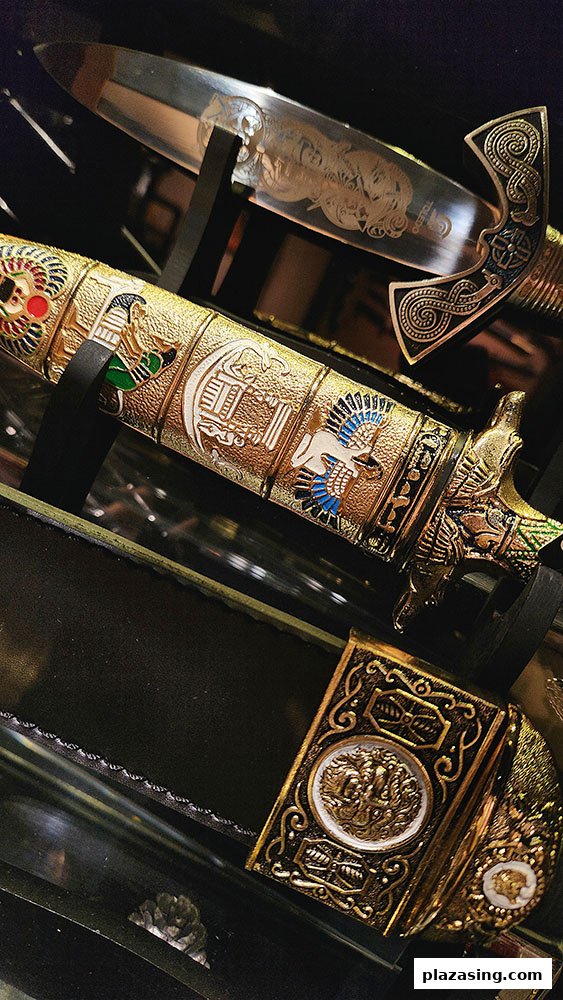 A close-up of a highly ornate, gold-colored decorative dagger with intricate historical and mythological designs.