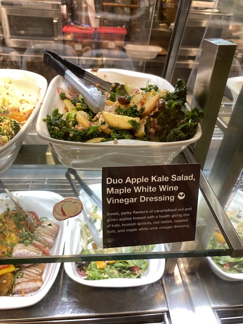 A vibrant Duo Apple Kale Salad with serving tongs in a display case at CafèMeal.