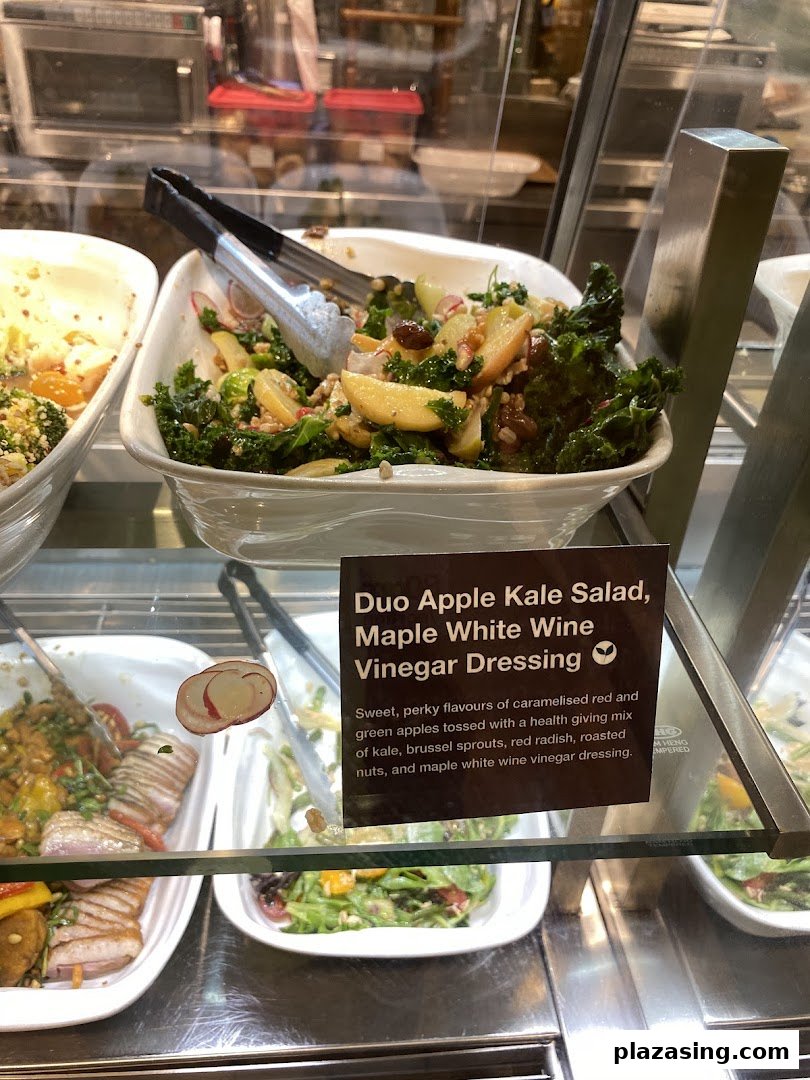A vibrant Duo Apple Kale Salad with serving tongs in a display case at CafèMeal.