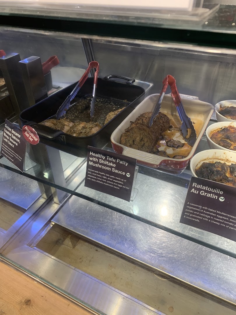 A deli counter showcasing prepared dishes like salmon, tofu patties, and ratatouille with descriptive labels.