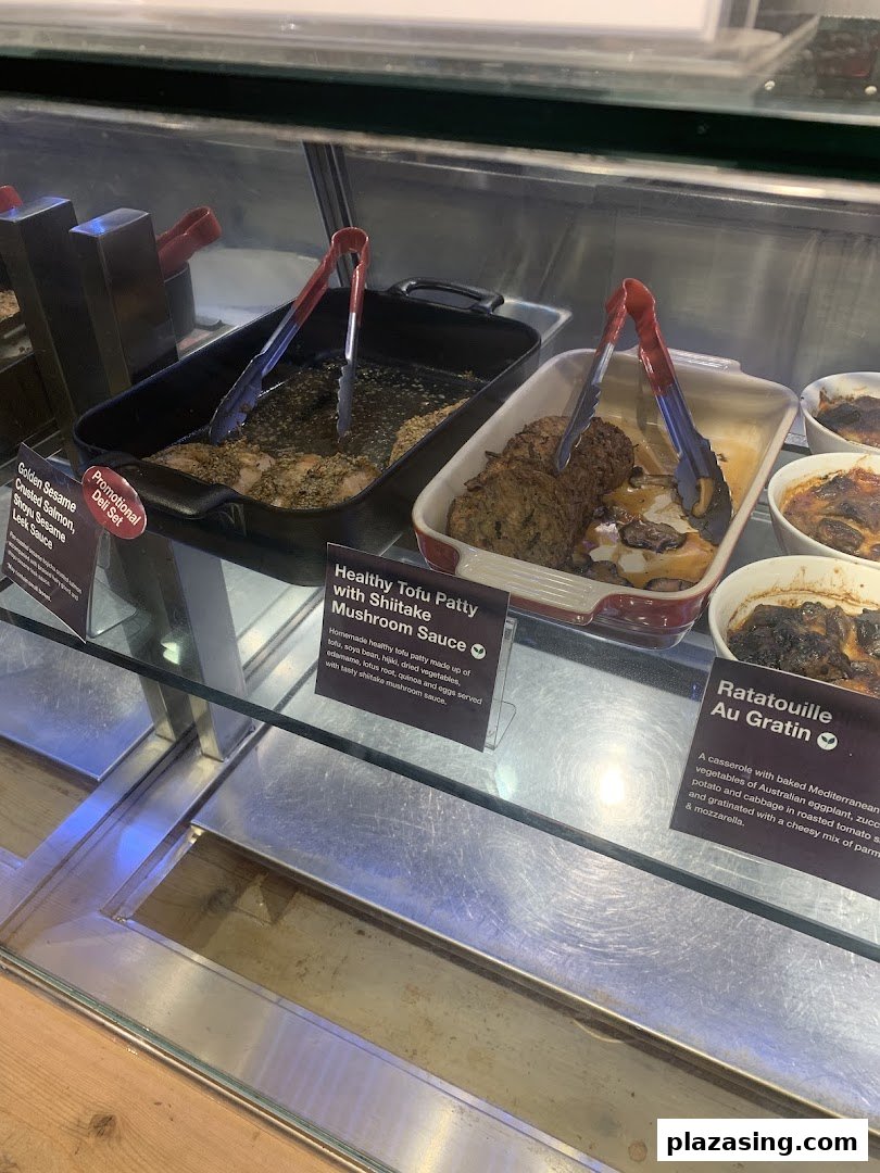A deli counter showcasing prepared dishes like salmon, tofu patties, and ratatouille with descriptive labels.