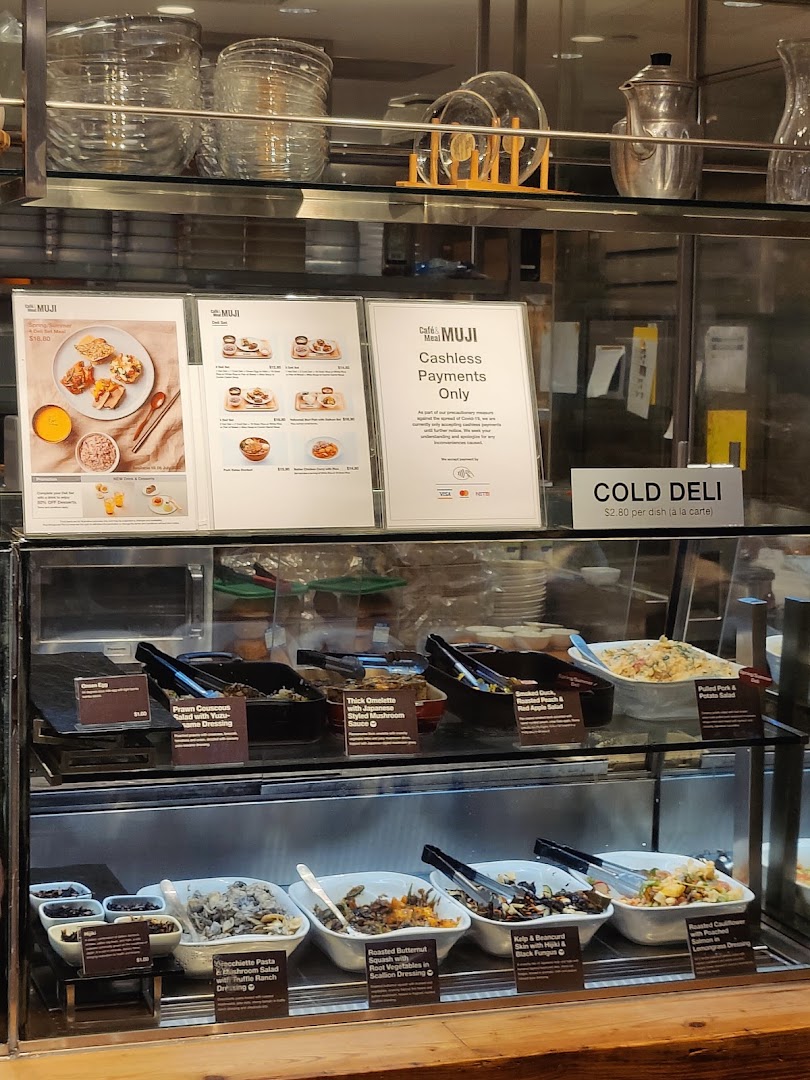 A display case at CafèMeal MUJI showcasing various cold deli dishes and menu boards for set meals.