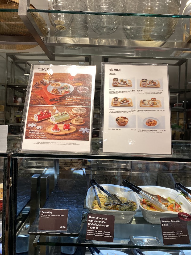 An interior view of CafèMeal MUJI, showcasing menu boards with festive specials and deli sets, alongside prepared food items.