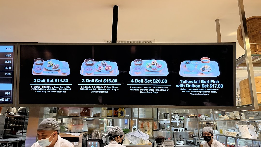 Digital menu board displaying deli sets and a fish dish, with kitchen staff preparing food below.