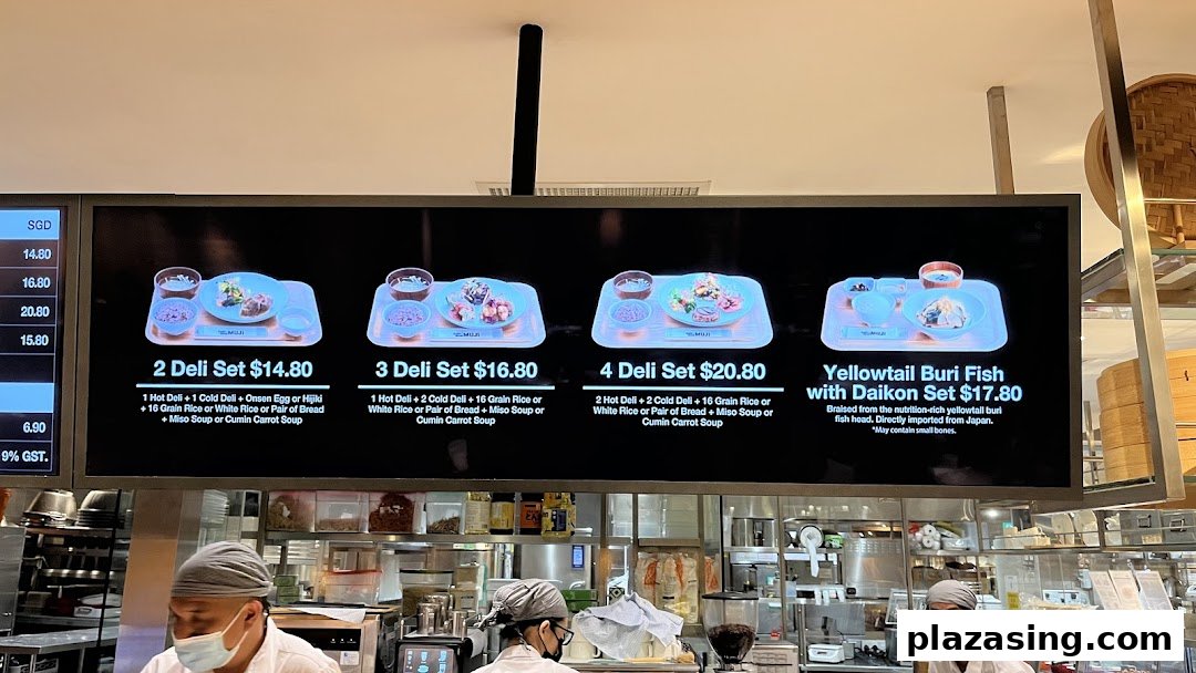 Digital menu board displaying deli sets and a fish dish, with kitchen staff preparing food below.