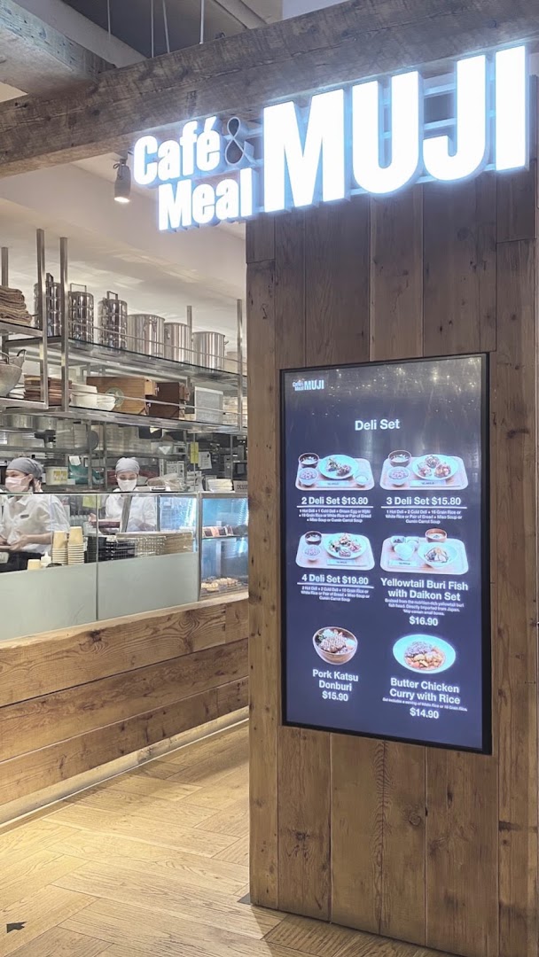 The entrance of Café & Meal MUJI featuring a digital menu board displaying various deli sets and main dishes.