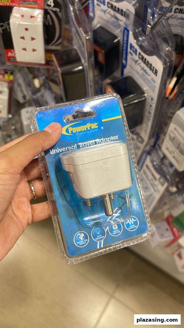 A hand holds a PowerPac universal travel adapter in its packaging, surrounded by other electronic accessories.