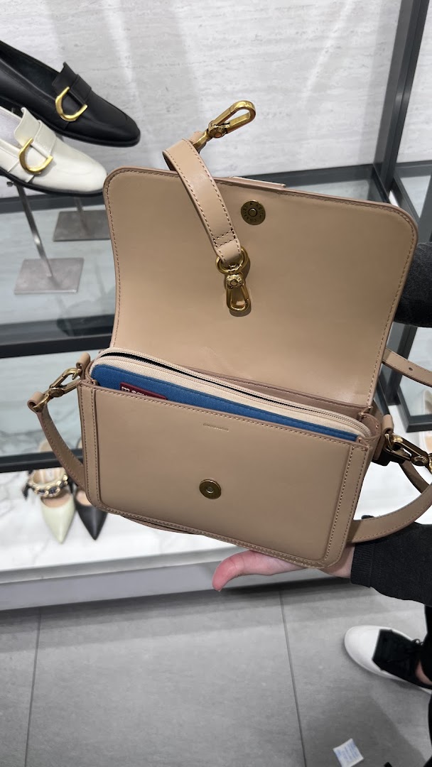 A person holds an open beige handbag, showcasing its interior and design, with shoes in the background.