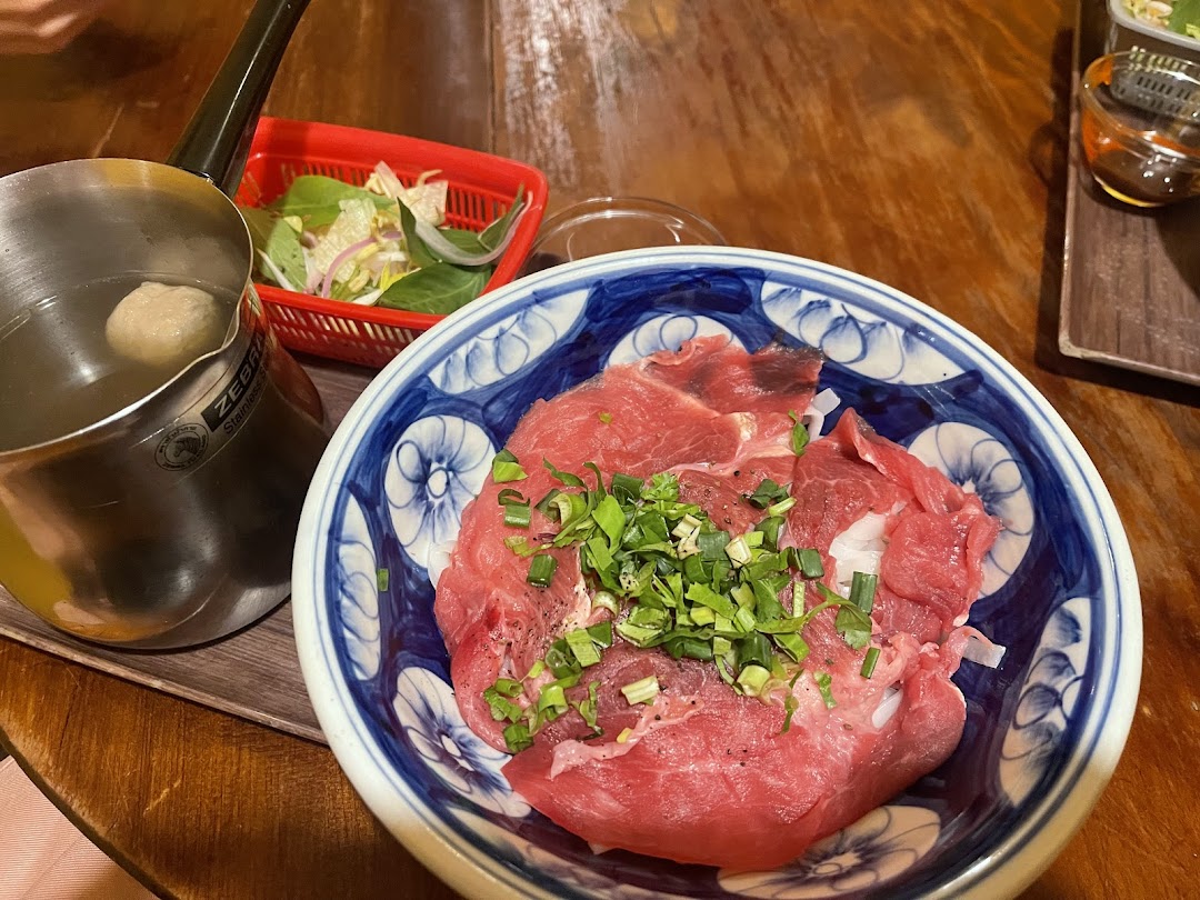 A bowl of raw beef with scallions, a pot of broth, and fresh greens on a wooden table.